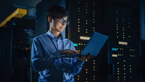 黑暗中的数据中心:男性it专家站在一排操作服务器机架旁,使用笔记本